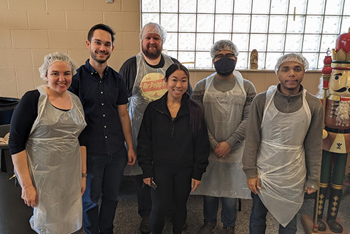 A group of smiling FASTies wear hair nets and aprons to serve meals in a homeless shelter