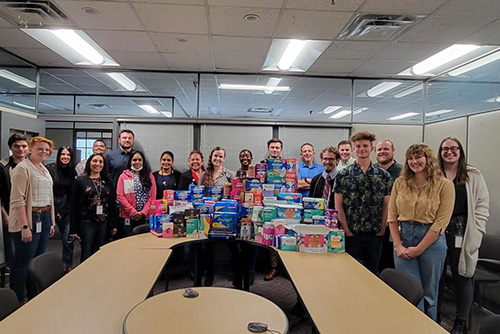 A group of smiling software employees stand around a desk filled with a large pile of hygiene products that they have collected for charity